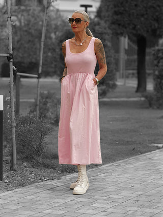 Frau trägt das OFF#DLY Kleid Dalia rosa, ein ärmelloses, knöchellanges Kleid mit geraffter Taille und Seitentaschen, kombiniert mit off-white Stiefeletten und goldfarbenen Accessoires.