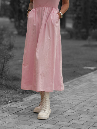 Person trägt das OFF#DLY Kleid Dalia rosa, ein fließendes, mid-calf Kleid mit Seitentaschen, kombiniert mit klobigen, cremefarbenen Stiefeln auf einem gepflasterten Weg.