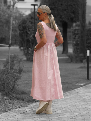 Person im OFF#DLY Kleid Dalia rosa, ärmelloses, knöchellanges Kleid mit fließendem Rockteil. Trägt graue Haare im Zopf, Tattoos, Sonnenbrille und helle Schuhe.