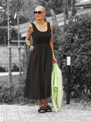 Eine reife Frau trägt das OFF#DLY Kleid Dalia black, ein ärmelloses, figurbetonendes Maxi-Kleid mit fließendem Rockteil, kombiniert mit schwarzen Sandalen und auffälligen Accessoires.