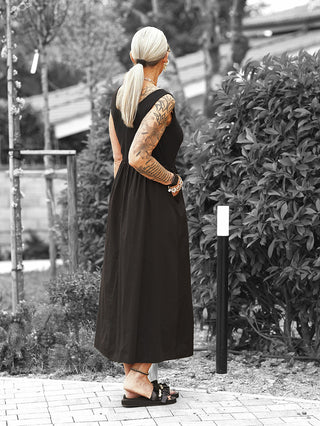 Person in ärmellosem OFF#DLY Kleid Dalia black, lang und locker fallend, mit schwarzem Materialmix, kombiniert mit schwarzen Sandalen und goldenen Accessoires, steht auf einem Weg mit Rücken zur Kamera.