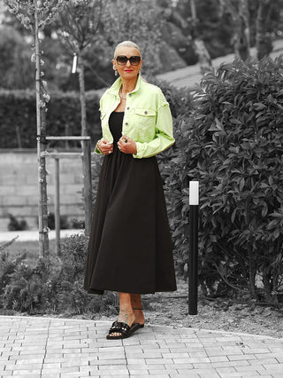 Ältere Frau trägt das OFF#DLY Kleid Dalia black, ein ärmelloses, fließendes Kleid mit feinem Rippenoberteil, betonter Taille und praktischen Seitentaschen, ergänzt durch eine gelbe Jeansjacke.
