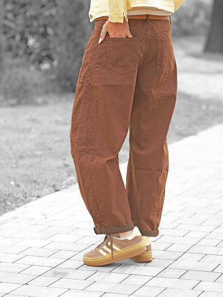 Person trägt OFF#DLY Barrel Color Denim Mia brown mit weitem Schnitt und umgeschlagenem Saum. Kombiniert mit Adidas Gazelle Sneakers, die Hand in der Gesäßtasche, zeigt der Look entspannten, modischen Stil.