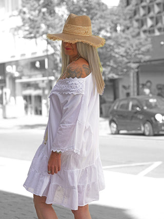 Frau in OFF#DLY Kleid Rabea white, einem luftigen, schulterfreien Sommerkleid mit Spitzenbesatz und Volants, trägt einen Strohhut, während sie auf einer sonnigen Stadtstraße steht.