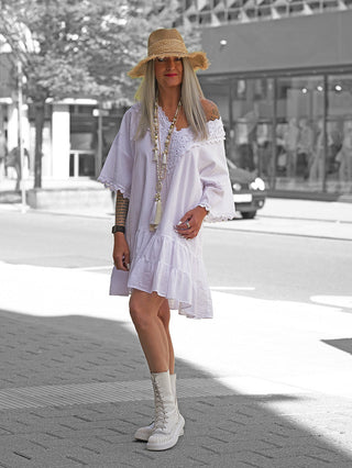 Frau trägt das OFF#DLY Kleid Rabea white: lockeres, weißes Sommerkleid mit Spitzenbesatz und Off-Shoulder-Ausschnitt. Kombiniert mit klobigen Stiefeln und Strohhut für einen lässigen Look.
