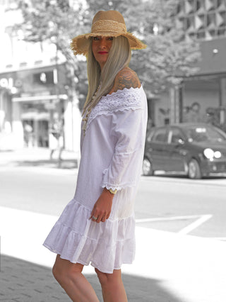 Person in lockerem OFF#DLY Kleid Rabea white mit schulterfreiem Ausschnitt und Spitzenbesatz, trägt einen Strohhut. Kleid hat Volants am Saum und vermittelt sommerliche Leichtigkeit.