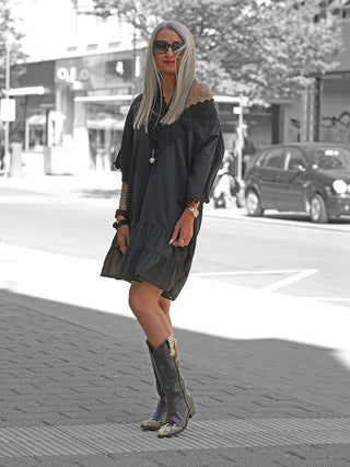 Frau trägt das OFF#DLY Kleid Rabea black, ein schwarzes, locker sitzendes Off-Shoulder-Sommerkleid mit Spitzenbesatz und Volants, kombiniert mit Accessoires und Cowboy-Stiefeln auf Stadtstraße.