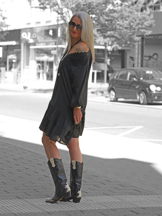 Frau in OFF#DLY Kleid Rabea black, einem schulterfreien schwarzen Sommerkleid mit Spitzenbesatz und Stufenrock, kombiniert mit kniehohen Cowboy-Stiefeln.