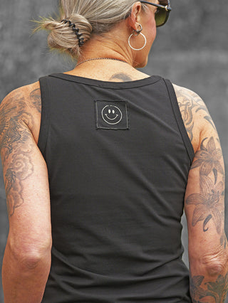 Person von hinten mit grauem Haarknoten, Sonnenbrille und Tattoos, trägt das OFF#DLY Top Nela schwarz mit weißem Smiley-Patch am Rücken. Schlichtes, ärmelloses Basic-Design.