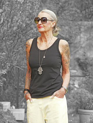 Frau trägt das OFF#DLY Top Nela black, ein figurbetontes, ärmelloses Oberteil aus glatter Baumwolle, kombiniert mit hellen Hosen; lässiger, eleganter Look mit Schmuck und Tattoos.