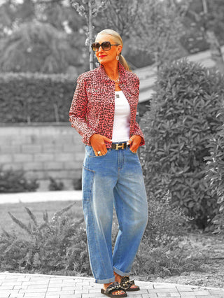 Frau in OFF#DLY Jacke Aice Leo Pink mit pink-schwarzem Leopardenmuster, über weißem Tanktop. Kombiniert mit blauen Jeans und Perlensandalen. Quadratische Sonnenbrille und Goldschmuck vervollständigen den Look.