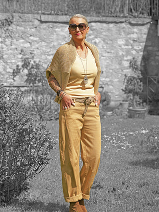 Ältere Frau trägt OFF#DLY Pant Senta camel, locker geschnittene Hose mit tiefen Taschen und Gürtel, kombiniert mit gelbem Top und Strickschal in einem Garten.