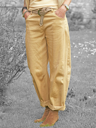 Person mit OFF#DLY Pant Senta camel in lockerem Schnitt, gelbe Hose mit gekrempelten Beinabschlüssen, Gürtel mit Nieten, getragen mit beigen Wildlederstiefeletten, Hände in vorderen Taschen.