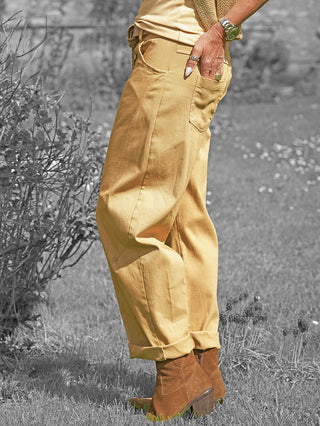 Person trägt OFF#DLY Pant Senta camel mit lockerem Schnitt und gekrempelten Bündchen, kombiniert mit braunen Wildlederstiefeletten, während die Hand mit Schmuck in die Fronttasche greift.