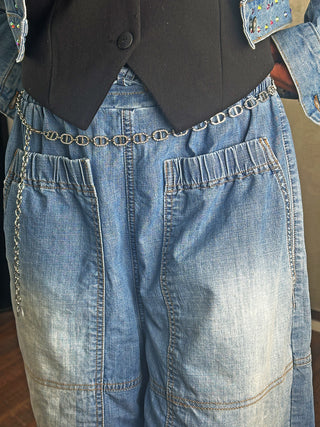 Nahaufnahme des OFF#DLY Kettengürtels silber, ein dekorativer Gürtel aus miteinander verbundenen ovalen Gliedern, der elegant über einem schwarzen, einreihigen Vest und luftigen, hellen Denim-Jeans getragen wird.