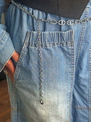 Nahaufnahme eines hellblauen Denim-Kleidungsstücks mit elastischen Taschen, das mit dem silbernen OFF#DLY Kettengürtel um die Taille getragen wird. Eine Hand steckt lässig in einer der Taschen.