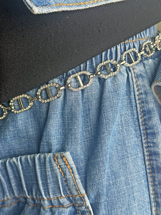 Nahaufnahme des OFF#DLY Kettengürtel silver, einem silbernen Kettengürtel mit ovalen, mit Strass besetzten Gliedern, drapiert über einem blauen Denim-Kleidungsstück mit sichtbarer Taschennaht und elastischem Bund.