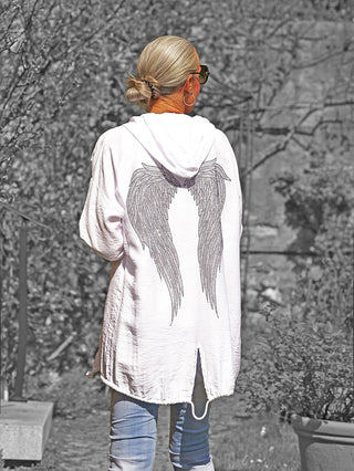 Person von hinten in einem Garten, trägt die OFF#DLY Jacke Samira white mit Kapuze und dekorativen Flügeln auf dem Rücken, kombiniert mit hellen Jeans, unterstreicht einen lässigen Look.