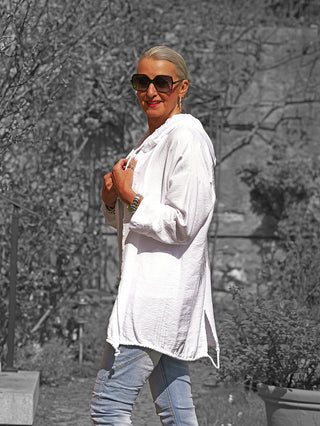 Erwachsener trägt OFF#DLY Jacke Samira white, eine locker fallende, weiße Hoodie-Jacke mit Kapuze, kombiniert mit Jeans. Die Person hält den Kragen, im Hintergrund sind Pflanzen sichtbar.