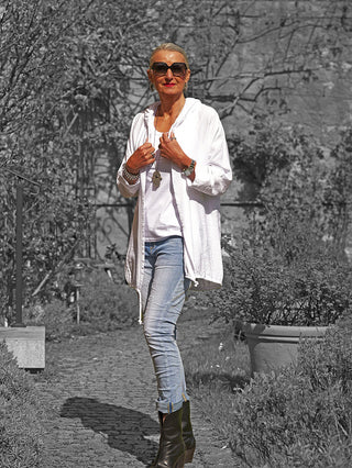 Frau in weißer OFF#DLY Jacke Samira mit Kapuze und lockerer Passform, zeigt Schmuck vor einem Gartenpfad. Ideal für Frühling/Sommer, sportlicher Look durch dekoratives Flügel-Design am Rücken.