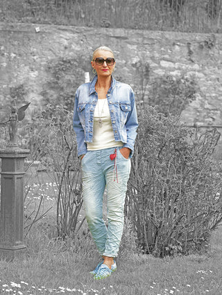 Frau trägt die OFF#DLY Jacke Sanna midblue, eine kurze, lässige Denim-Jacke mit Knopfleiste und Brusttaschen, kombiniert mit einer weißen Bluse, in einem Garten stehend.