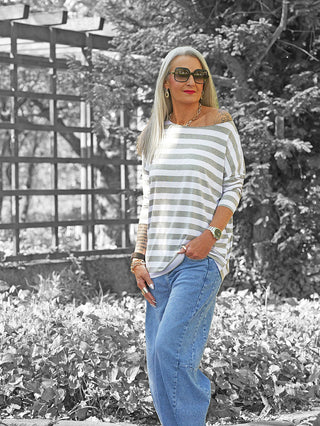 Frau mit silbernem Haar trägt das OFF#DLY Ringel Grosso grau/weiß gestreifte Langarmshirt, kombiniert mit blauer Jeans und auffälligen Accessoires, vor einem Laub-Hintergrund.