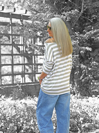 Frau im Garten trägt OFF#DLY Ringel Grosso grey/white, ein locker geschnittenes, gestreiftes Langarmshirt. Sie zeigt ihr Profil, während sie über die Schulter schaut, mit schulterlangem Haar und Tattoo sichtbar.