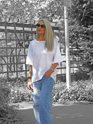 Frau in OFF#DLY Shirt Rieke weiß, locker geschnitten, aus Leinen und Baumwolle, mit langen Ärmeln, kombiniert mit lässigen blauen Jeans und goldenen Accessoires, spaziert draußen.