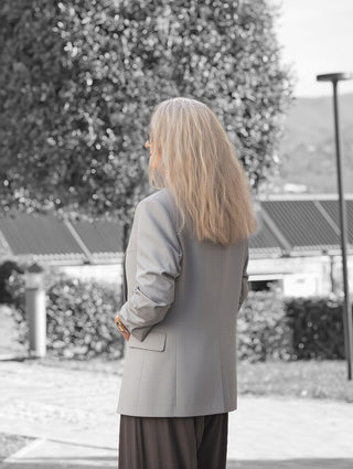 Eine Person trägt den OFF#DLY Blazer Dana grey mit klassischem Reverskragen und aufgesetzten Taschen, stehend in einem landschaftlich gestalteten Bereich.