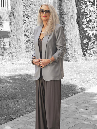 Erwachsene Frau trägt den OFF#DLY Blazer Dana grey mit klassischem Reverskragen und aufgesetzten Taschen, kombiniert mit einem schwarzen Spitzentop und weiten dunklen Hosen in einem parkähnlichen Umfeld.