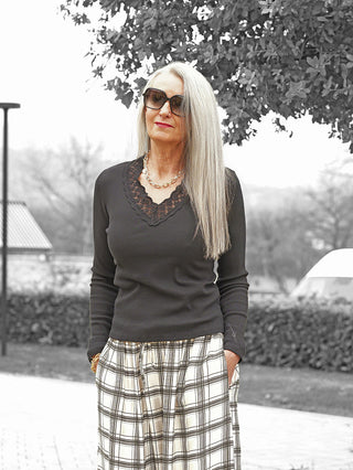 Eine reife Frau trägt das OFF#DLY Longsleeve Enie black mit Spitzen-V-Ausschnitt, kombiniert mit einer karierten Hose, und posiert elegant mit goldfarbenem Schmuck auf einem gepflasterten Weg.