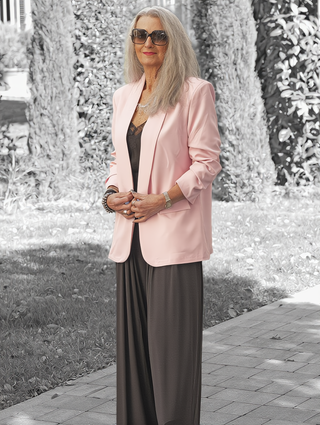 Eine reife Frau trägt den OFF#DLY Blazer Dana rose, einen offenen, leicht rosa Blazer mit klassischem Revers. Der Blazer ergänzt ihr schwarzes Spitzenoberteil und weite Hosen.