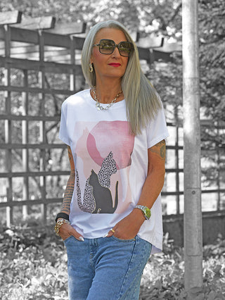 Frau mit grauen Haaren trägt das OFF#DLY Tee Cat´s white/pink, ein weißes T-Shirt mit Katzenmotiv, locker in blaue Jeans gesteckt, kombiniert mit Sonnenbrille und Schmuck, vor Gartenhintergrund.