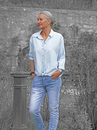 Eine ältere Frau trägt die OFF#DLY Bluse Sandra Stripes lightblue/white, ein leichtes, fließendes Hemd mit Streifen, locker gebunden und kombiniert mit einer Jeans, ideal für warme Tage.