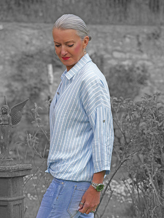 Frau in einem Garten trägt die OFF#DLY Bluse Sandra Stripes lightblue/white, ein leichtes, fließendes Hemd mit feinem Streifenmuster, hochgekrempelten Ärmeln und eleganter, zeitloser Leichtigkeit.