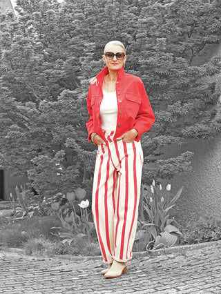 Frau in einem Garten trägt die OFF#DLY Jacke Modica red mit offenem Kragen, kombiniert mit einer weißen Tanktop und gestreiften Hosen. Accessoires: Sonnenbrille, Creolen, Armbänder.