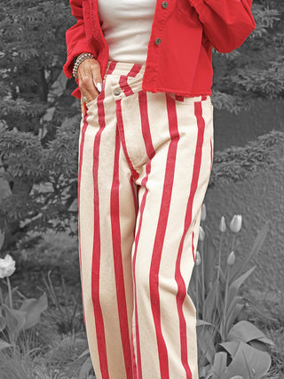 Person trägt Lumina Hose Davina beige/rot, auffällig gestreifte, hoch taillierte Hose. Rote Jacke, weiße Tanktop, Hand mit Schmuck in Hosentasche. Hintergrund: Tulpen und Laub.