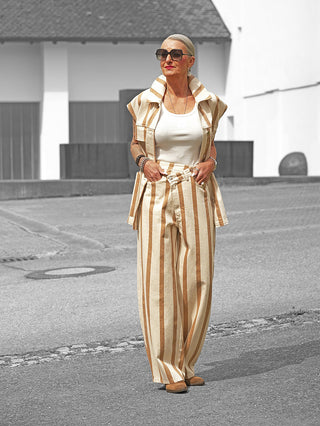Frau in Lumina Hose Davina beige/camel mit weiten Beinen und stilvollem Streifenmuster, kombiniert mit einem tanktop und einem offenen, ärmellosen Vest, spaziert selbstbewusst vor modernem Gebäude.