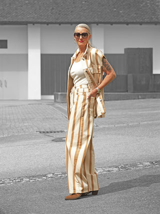 Frau trägt Lumina Hose Davina beige/camel mit breitem Streifenmuster und passender ärmelloser Weste. Moderne, lässige Silhouette mit auffälliger Knopflösung. Kombiniert mit weißem Tanktop und braunen Stiefeletten.