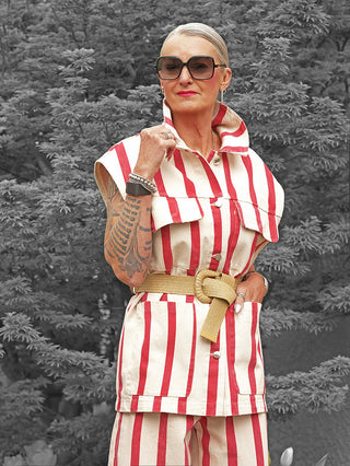 Frau in Lumina Gilet Delia beige/rot mit auffälligem Streifenmuster, kombiniert mit passender Hose, breitem Gürtel und großem Kragen, trägt große Sonnenbrille, Schmuck und hat eine tätowierte Hand.