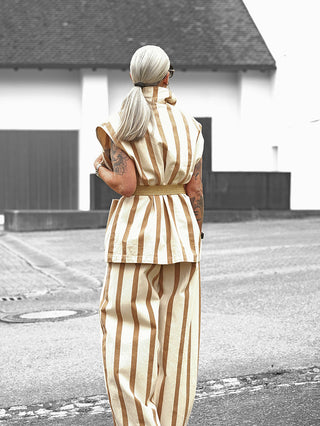Person von hinten, trägt Lumina Gilet Delia beige/camel mit vertikalen Streifen, ärmelloser Schnitt, Bindegürtel. Kombiniert mit passender Hose, breite Beine. Tattoos sichtbar auf Unterarmen, vor weißem Gebäude.
