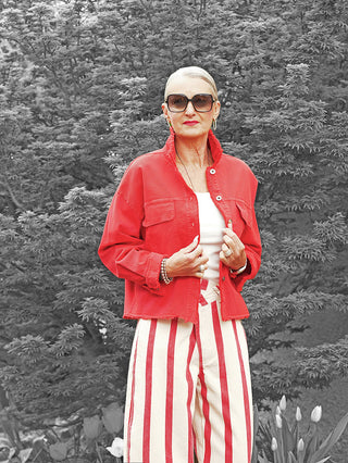 Frau trägt die OFF#DLY Jacke Modica red, eine kurze rote Jacke mit offenen Saumkanten und Brusttaschen, über einem weißen Tanktop, kombiniert mit gestreiften Hosen und Sonnenbrille.