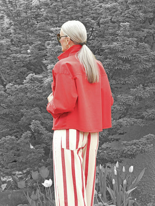 Eine Person trägt die OFF#DLY Jacke Modica red, mit kurzem, modischem Schnitt und offenen Abschlüssen, kombiniert mit hochtaillierten gestreiften Hosen.