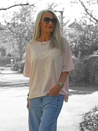 Frau trägt OFF#DLY Sweat Level 1 rosa, ein leichtes, lockeres Sweatshirt mit modernem Print, kombiniert mit blauer Jeans und goldfarbenem Schmuck, während sie auf einem baumgesäumten Weg geht.
