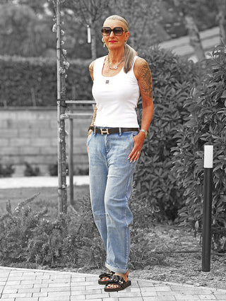 Frau trägt OFF#DLY Denim Bea midblue: Relaxed-Fit Jeans mit umgeschlagenen Säumen, klassischem Five-Pocket-Stil und schwarzem Gürtel. Kombiniert mit weißem Top und schwarzen Sandalen mit Perlen.