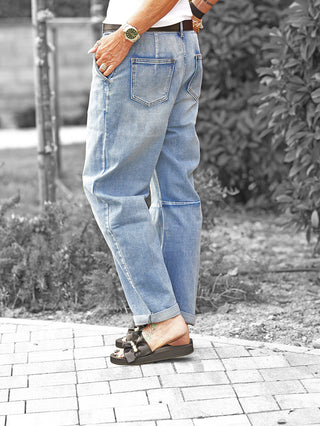 Person trägt die OFF#DLY Denim Bea midblue, locker geschnittene, hell verwaschene Jeans, umgeschlagener Saum, Hand in Gesäßtasche, kombiniert mit schwarzen Schleifensandalen auf einem gepflasterten Weg.