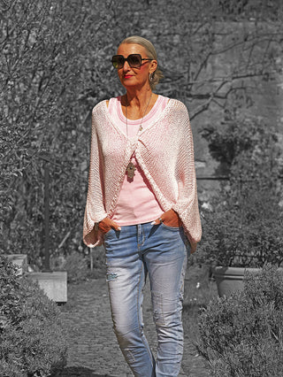 Ältere Frau trägt OFF#DLY Netzoberteil Klara rosa, lässig über rosa T-Shirt, kombiniert mit Jeans und Sonnenbrille, in einem Gartenbereich mit Pflanzen und Hintergrund aus Mauerwerk.