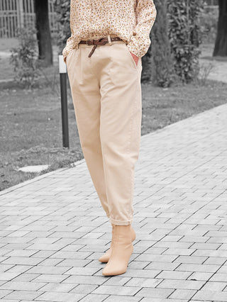 Frau trägt OFF#DLY Hose Amy beige: hochgeschnittene, plissierte Chinos mit dünnem braunen Gürtel, kombiniert mit floraler Bluse und nudefarbenen Stiefeletten, Hände lässig in den Taschen.