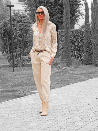 Frau trägt die OFF#DLY Hose Amy beige, locker geschnitten mit elastischem Bund und schmalem Gürtel, kombiniert mit einer floralen Bluse und beigen Stiefeletten auf einem gepflasterten Weg.