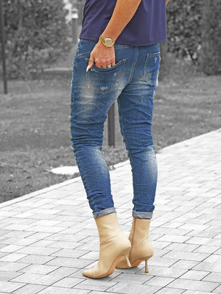 Person trägt OFF#DLY Denim Detroy Alassio blue, schmal geschnittene, leicht zerstörte blaue Jeans, an den Knöcheln umgeschlagen. Eine Hand mit Goldarmbanduhr steckt lässig in der Gesäßtasche.